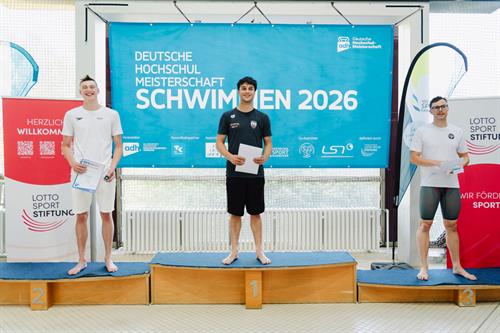 Drei Schwimmer stehen bei der Deutschen Hochschulmeisterschaft Schwimmen 2026 auf dem Siegerpodest. Der Erstplatzierte steht mittig auf dem Podest und hält seine Urkunde in der Hand.